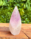 Teardrop-shaped crystal with pink and purple hues on a wooden surface with a green foliage background
