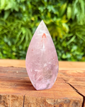 Teardrop-shaped pink crystal on a wooden surface with a green leafy background