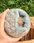Hand holding a large crystal celestite freeform with a natural pattern against a green foliage background