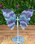 Butterfly-shaped decorative item with purple and blue wings on a wooden surface with greenery in the background