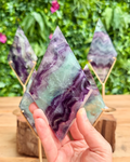 Hand holding a tdiamond shaped fluorite crystal with purple and green patterns against a blurred green background