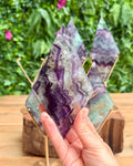 Hand holding a purple crystal  diamond with more crystals in the background on a wooden stand.