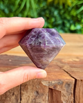 Hand holding a purple crystal on a wooden surface with greenery in the background