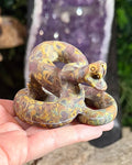 A hand holding a Ajooba jasper coiled snake carving against a blurred background.