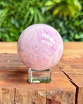 Pink kunzite crystal sphere on a wooden surface with green foliage in the background