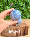 Hand holding a blue kyanite sphere with another sphere in the background on a wooden surface.