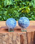 Two blue kyanite crystal spheres on a wooden surface with a green foliage background