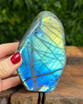 Multicolored labradorite stone held by a hand with red nail polish on a wooden surface.