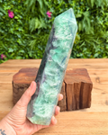 Hand holding a large green crystal on a wooden surface with a blurred green foliage background