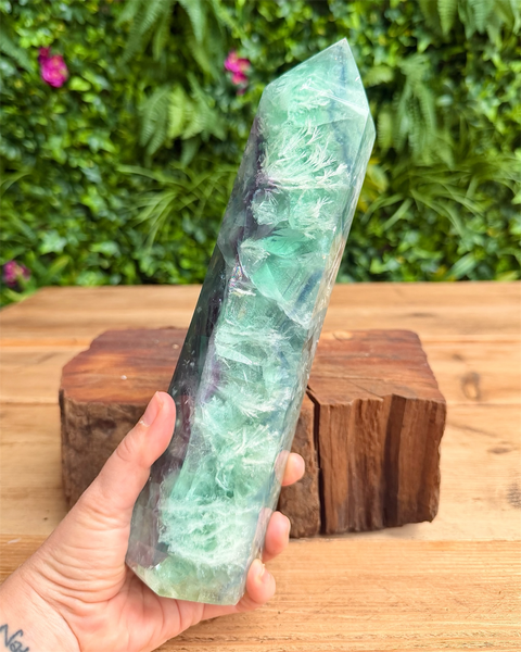 Hand holding a large green crystal on a wooden surface with a blurred green foliage background