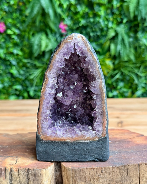 A carved amethyst geode sculpture with a dark gray base, displayed on a rustic wooden surface with a blurred green foliage background.