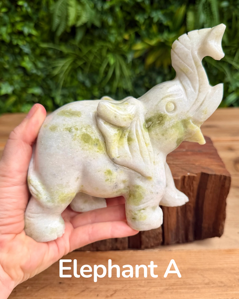 Hand holding a stone elephant figurine with a natural greenish patina, set against a blurred green foliage background.