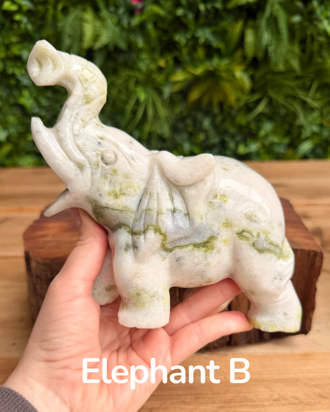 Hand holding a white jade elephant sculpture with green veins against a blurred green foliage background.