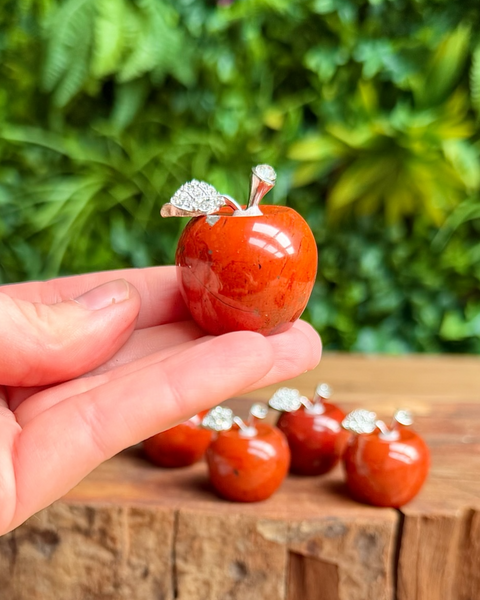 Hand holding a red apple-shaped red jasper crystal with a diamond-like top, set against a blurred green outdoor background.