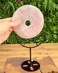 Rose quartz donut stone on a black stand with a hand holding it, against a green foliage background.