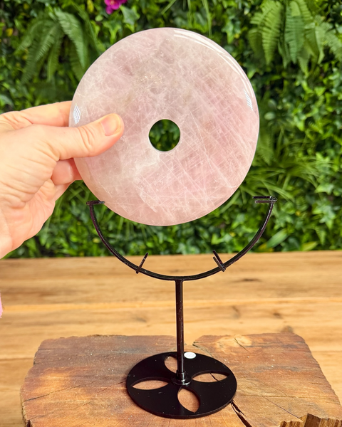 Rose quartz donut stone on a black stand with a hand holding it, against a green foliage background.