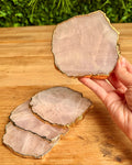 A hand holds a polished rose quartz lice coaster showing a pale pink stone face and a metallic gold leaf rim, with two more coasters stacked on a wooden tabletop.
