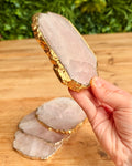 A hand holds a polished pale pink rose quartz coaster  slice with a shiny gold leaf edge, with two more slices resting on a wooden surface below against a blurred green foliage background.