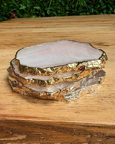 Three stacked natural crystal or stone coasters with irregular polished edges and metallic gold leaf detailing, displayed on a wooden tabletop with green foliage in the background.