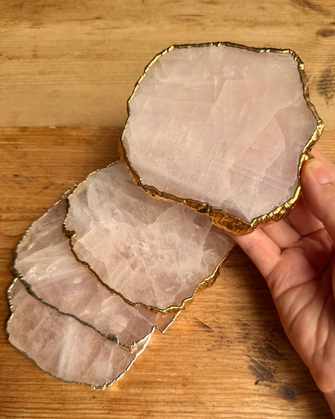 Four rose quartz crystal coasters with gold-rimmed edges, with one held by a hand over a wooden surface.