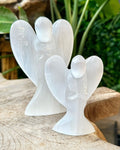 Two selenite crystal angel figures of different sizes placed on a wooden surface with foliage in the background.
