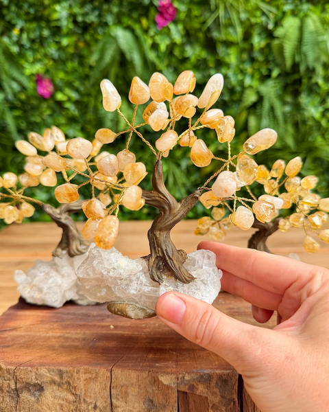 A hand holding a crystal tree of hope ornament made of yellow quartz crystals on a copper wire, set on a clear quartz crystal cluster base, with two more trees in the background against a green foliage backdrop.