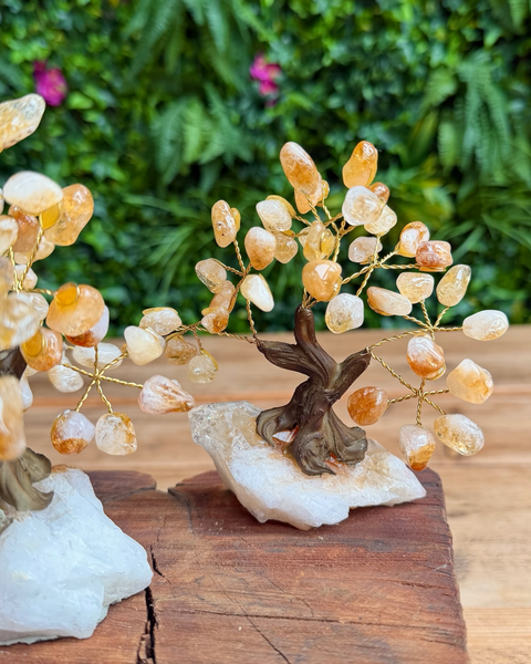A crystal tree of life figurine made of small amber and clear quartz stones on gold-toned wire branches, mounted on a white raw stone base and displayed on a wooden surface with a blurred green garden background.