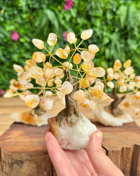 A hand holding a small crystal tree of light amber and yellow quartz with a brown wire trunk and a white stone base, with two more trees blurred in the background on a wooden surface and green foliage.
