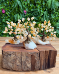 Three small crystal tree figurines with amber and cream-colored gemstone leaves on brown wire trunks, displayed on a rustic wooden log slice with a blurred green foliage background.