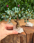 A hand holding a crystal tree of life sculpture made of green stone leaves on gold wire branches, mounted on a clear quartz crystal base, set on a rustic wooden surface with a blurred green garden background.