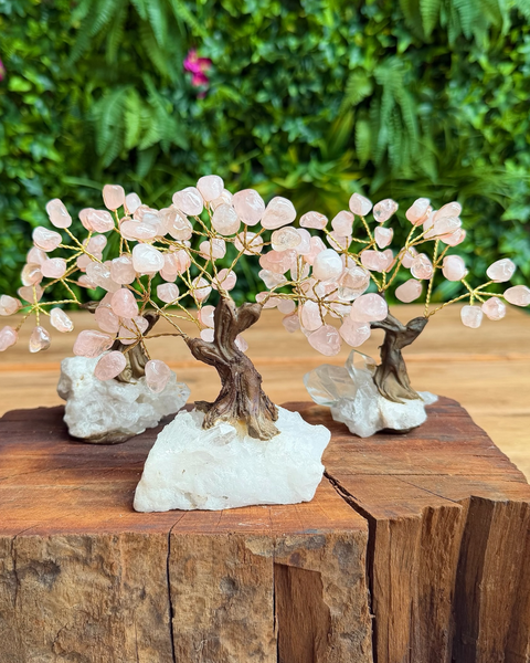 Three rose quartz crystal tree mini sculptures with gold wire branches and pink crystal leaves, mounted on white crystal bases and displayed on a rustic wooden block with a green foliage background.