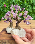A small crystal tree of life figurine with purple amethyst leaves and a brown wire trunk, mounted on a clear quartz base and held in a person's hand against a blurred green outdoor background.