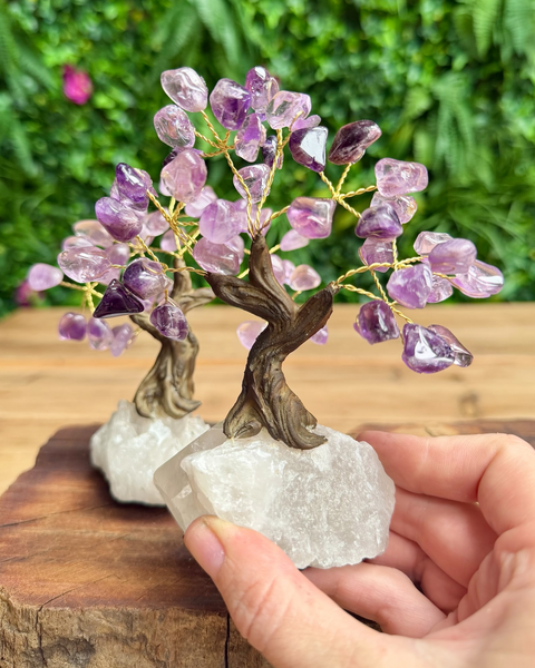 A small crystal tree of life figurine with purple amethyst leaves and a brown wire trunk, mounted on a clear quartz base and held in a person's hand against a blurred green outdoor background.