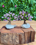 Two miniature crystal tree figurines with purple amethyst blossoms and brown branches, mounted on light gray stone bases and displayed on a rustic wooden log with a green foliage background.