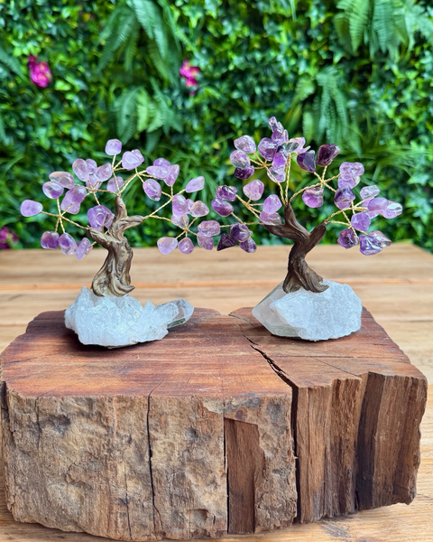 Two miniature crystal tree figurines with purple amethyst blossoms and brown branches, mounted on light gray stone bases and displayed on a rustic wooden log with a green foliage background.