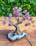 A crystal tree of life figurine made of purple amethyst stones and gold wire branches, mounted on a twisted brown tree trunk and a blue-gray geode base, displayed on a wooden surface with a blurred green foliage background.