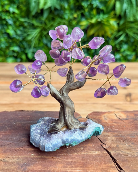 A crystal tree of life figurine made of purple amethyst stones and gold wire branches, mounted on a twisted brown tree trunk and a blue-gray geode base, displayed on a wooden surface with a blurred green foliage background.