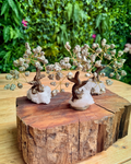 Three miniature crystal tree ornaments with gold wire branches, dried white flower clusters, and brown faux-wood trunks, displayed on a rustic wood slice pedestal with a blurred green garden background.