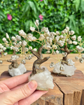 A hand holds a small metallic crystal tree ornament with silver-toned branches and clear quartz crystal bases, with two more trees in the background on a wooden surface and a blurred green garden backdrop.