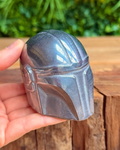 Metallic helmet resembling a character from a popular franchise held in a hand with a wooden surface and greenery in the background.