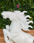 White jade horse sculpture with a flowing mane on a wooden surface with a green leafy background.