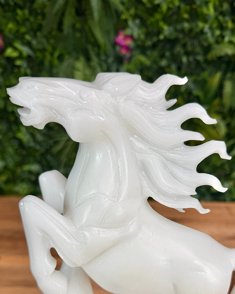 White jade horse sculpture with a flowing mane on a wooden surface with a green leafy background.