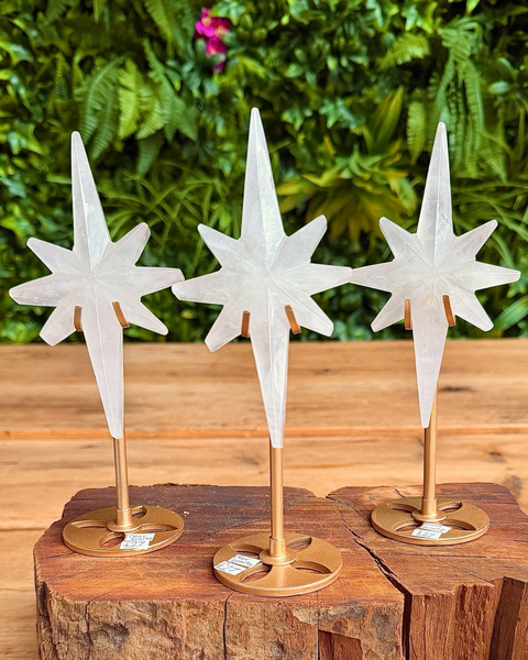 Three white star-shaped clear quartz stars on gold stands placed on a wooden block with a green leafy background.