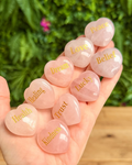 Hand holding pink heart-shaped stones with gold inscriptions against a blurred green background