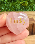 Heart-shaped rose quartz stone with 'Lucky' engraved, held in hand with greenery in the background