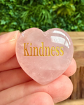 Heart-shaped rose quartz stone with 'Kindness' engraved, held in hand with greenery in the background