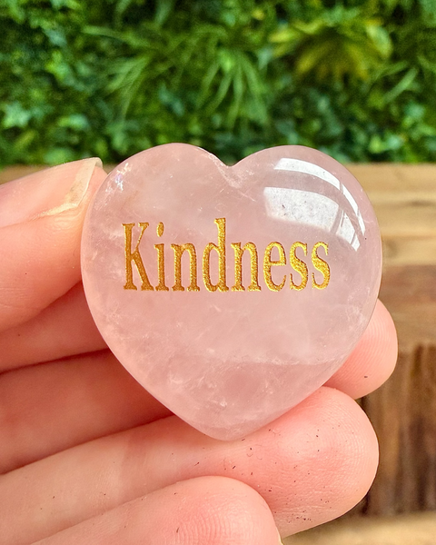 Heart-shaped rose quartz stone with 'Kindness' engraved, held in hand with greenery in the background