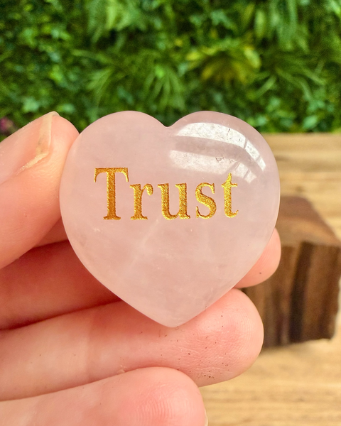 Heart-shaped rose quartz crystal with 'Trust' engraved, held in hand against a blurred natural background.