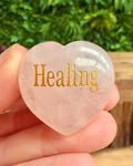 Heart-shaped crystal with 'Healing' engraved on it held in a hand against a blurred green background.