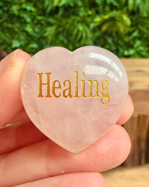 Heart-shaped crystal with 'Healing' engraved on it held in a hand against a blurred green background.
